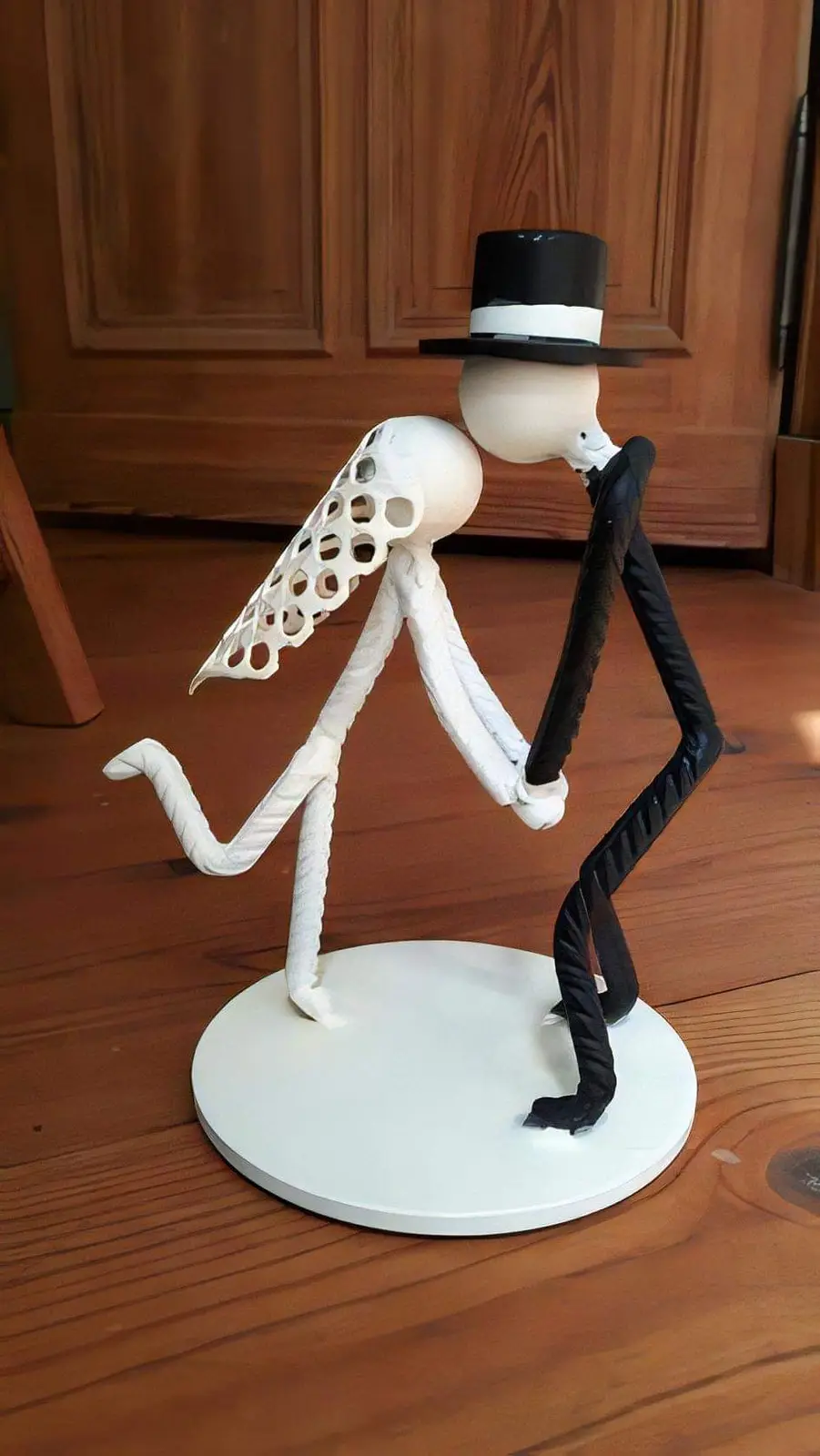 A handmade figurine depicts two stick-figure-like characters dancing on a white circular base. One figure is white with a…