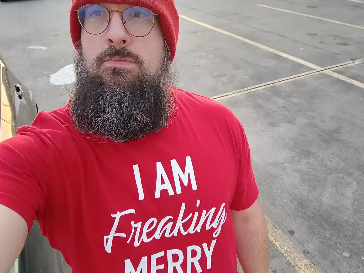 A man with a full beard, wearing a red beanie and glasses, takes a selfie in a parking lot while wearing a red t-shirt that…