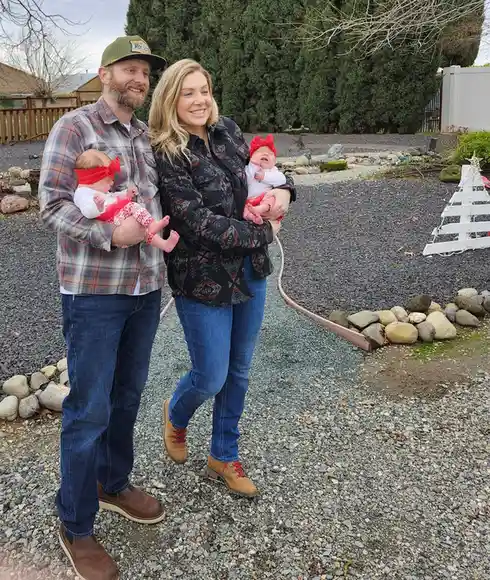 A smiling man and woman stand outdoors on a gravel path, each holding a baby dressed in red headbands and festive outfits…