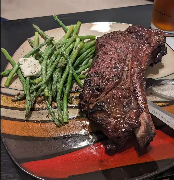 A close-up photo of a large, charred steak with a bone, served alongside a pile of green asparagus spears topped with a pat…