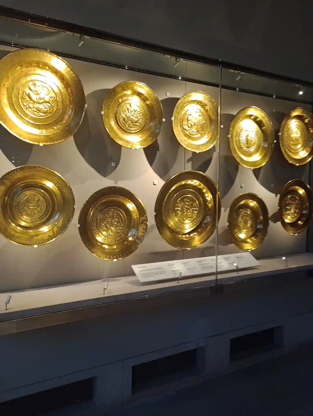 A museum display features nine ornate, golden, circular plates mounted on a wall behind glass. Each plate has intricate…