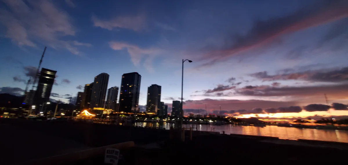A blurry photo of a city skyline at dusk with tall buildings, a waterfront, and a colorful sunset sky. A small white sign in…