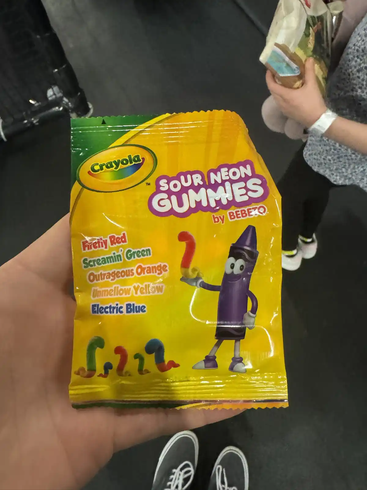 A hand holds a yellow Crayola Sour Neon Gummies bag featuring a purple crayon mascot and colorful gummy worm illustrations…