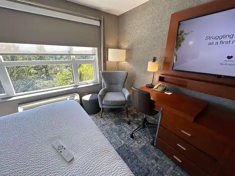 A hotel room with a bed, desk, chair, and large window showing trees. A TV mounted on the wall displays the text 'Struggling…