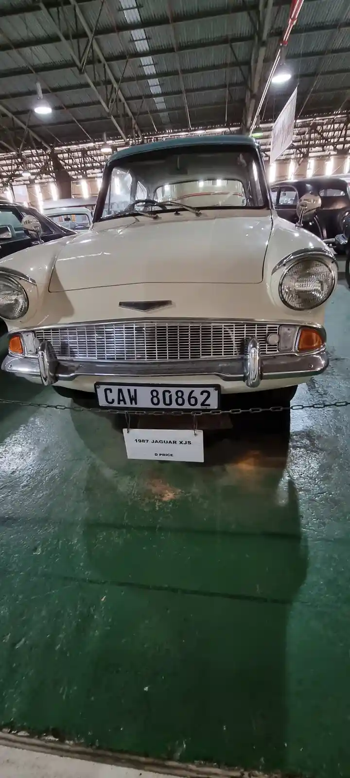 A vintage cream-colored car with a blue roof is displayed indoors on a green floor, its front grille and round headlights…