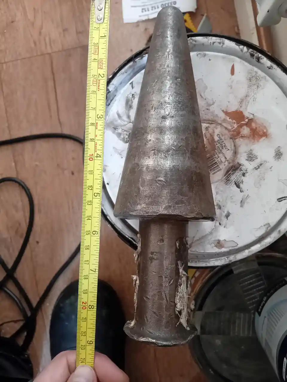 A hand holds a yellow Stanley tape measure next to a large, worn, metallic cone-shaped object, which appears to be a tool or…