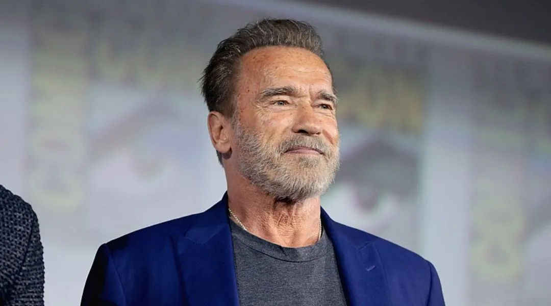 arnold schwarzenegger is shown with a smug, self-satisfied smirk while wearing a blue blazer over a gray t-shirt. The…