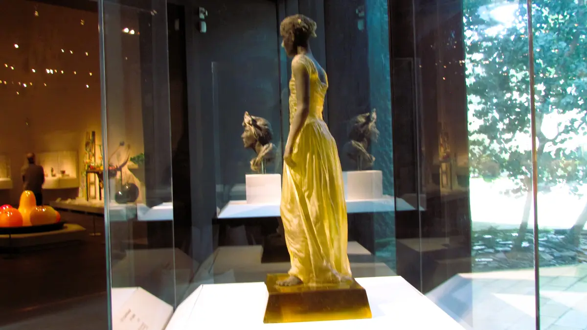 A full-length statue of a woman in a flowing yellow dress stands inside a glass display case in a museum. Behind her, two…