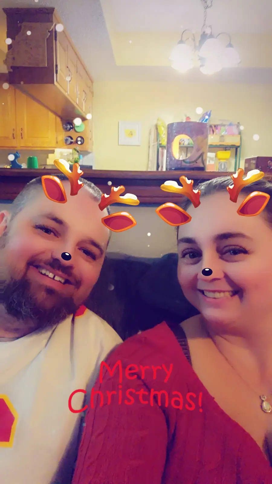 A selfie of a smiling man and woman indoors, both with cartoon reindeer antlers and black noses superimposed on their faces…