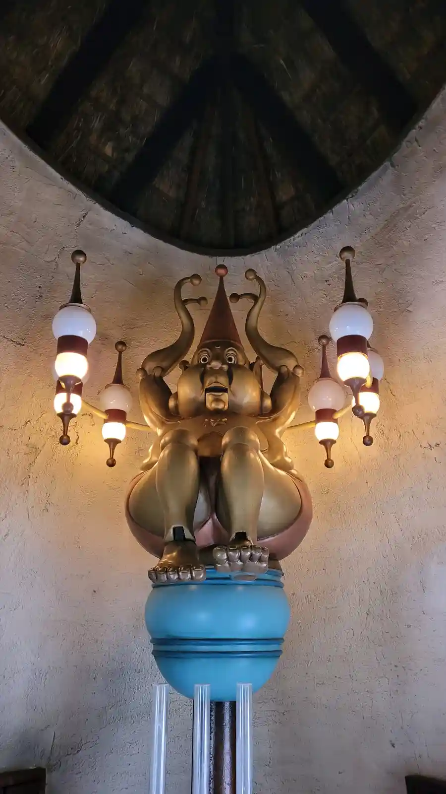 A golden statue of a jester with a surprised expression sits atop a blue pedestal, surrounded by lit lamps. The overlaid…
