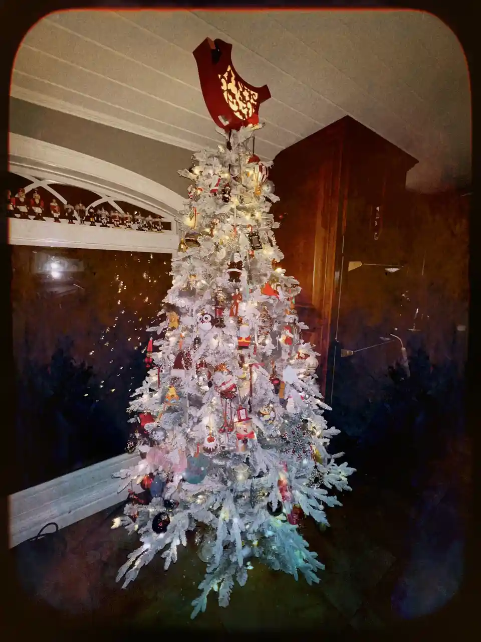 A heavily decorated white flocked Christmas tree with numerous ornaments, lights, and a red bird-shaped topper stands in a…