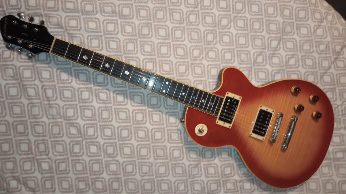A red and orange sunburst electric guitar with a black fretboard and white cross-shaped inlays lies diagonally on a white…