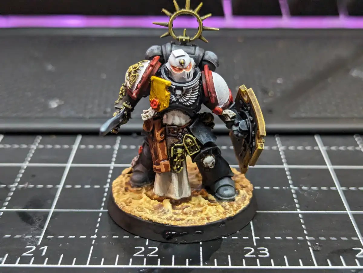 A painted miniature of a Space Marine from Warhammer 40,000, wearing white and black armor with gold accents, holding a…