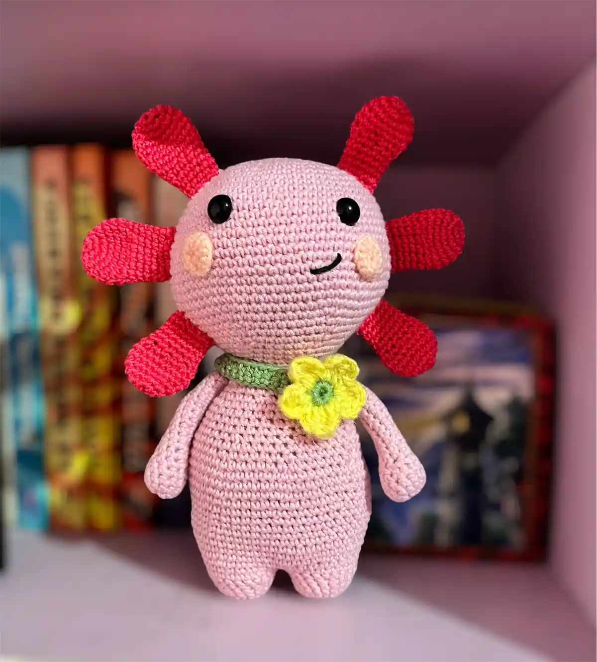 A pink crocheted plush toy with red appendages and a yellow flower on its green collar sits on a shelf, with books and a…