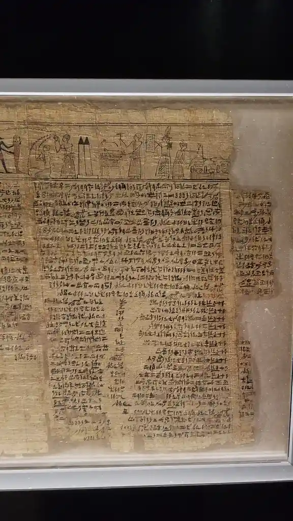An ancient Egyptian papyrus scroll, displayed behind glass, features hieroglyphic text and a top row of stylized human…