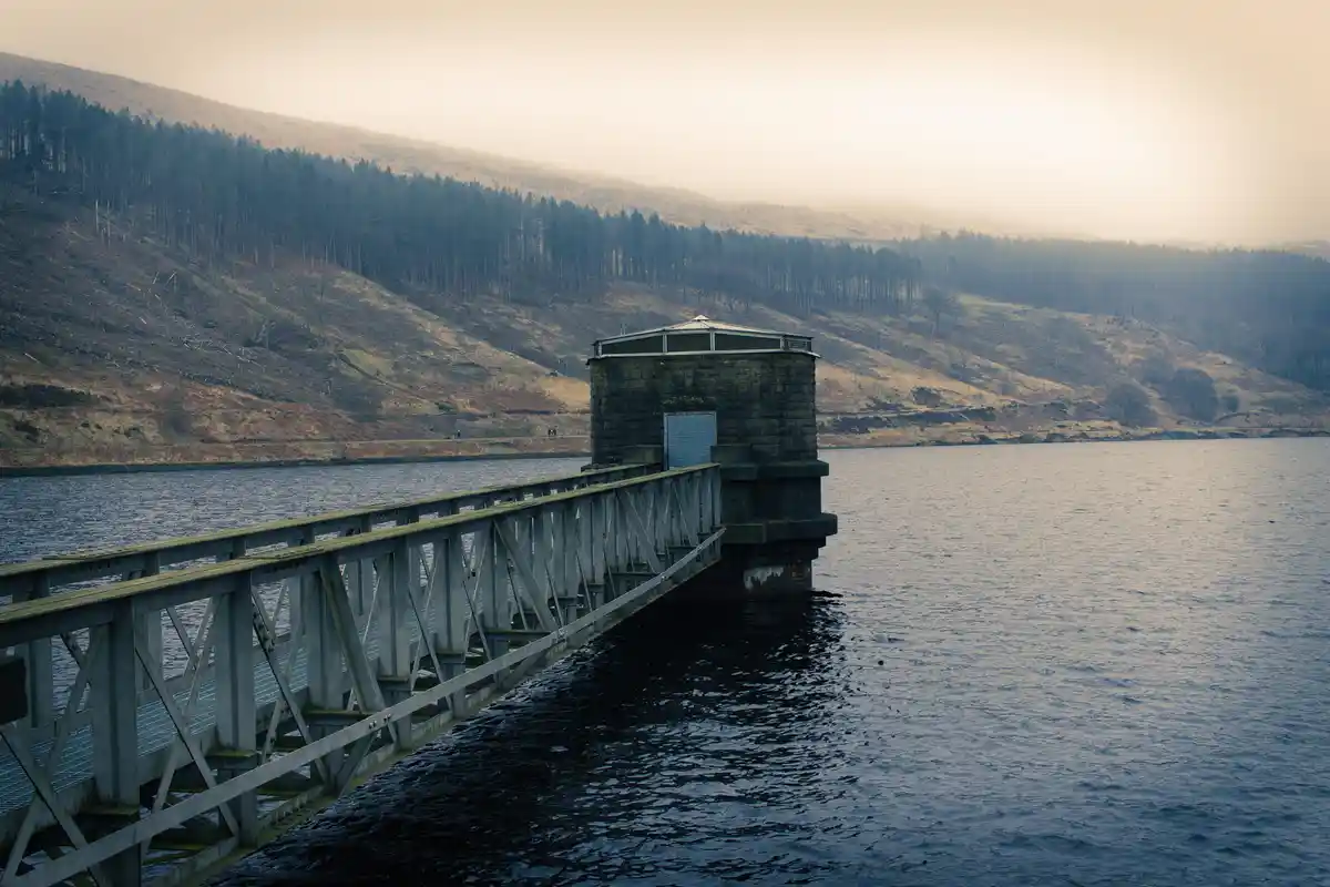 A serene, misty landscape featuring a metal walkway extending from the shore to a stone tower structure on a calm lake, with…