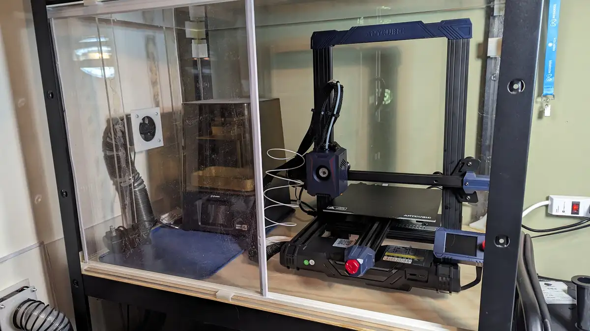 A clear enclosure houses two 3d printers side by side: an Anycubic model on the right and a Phrozen Sonic Mini 4k on the…