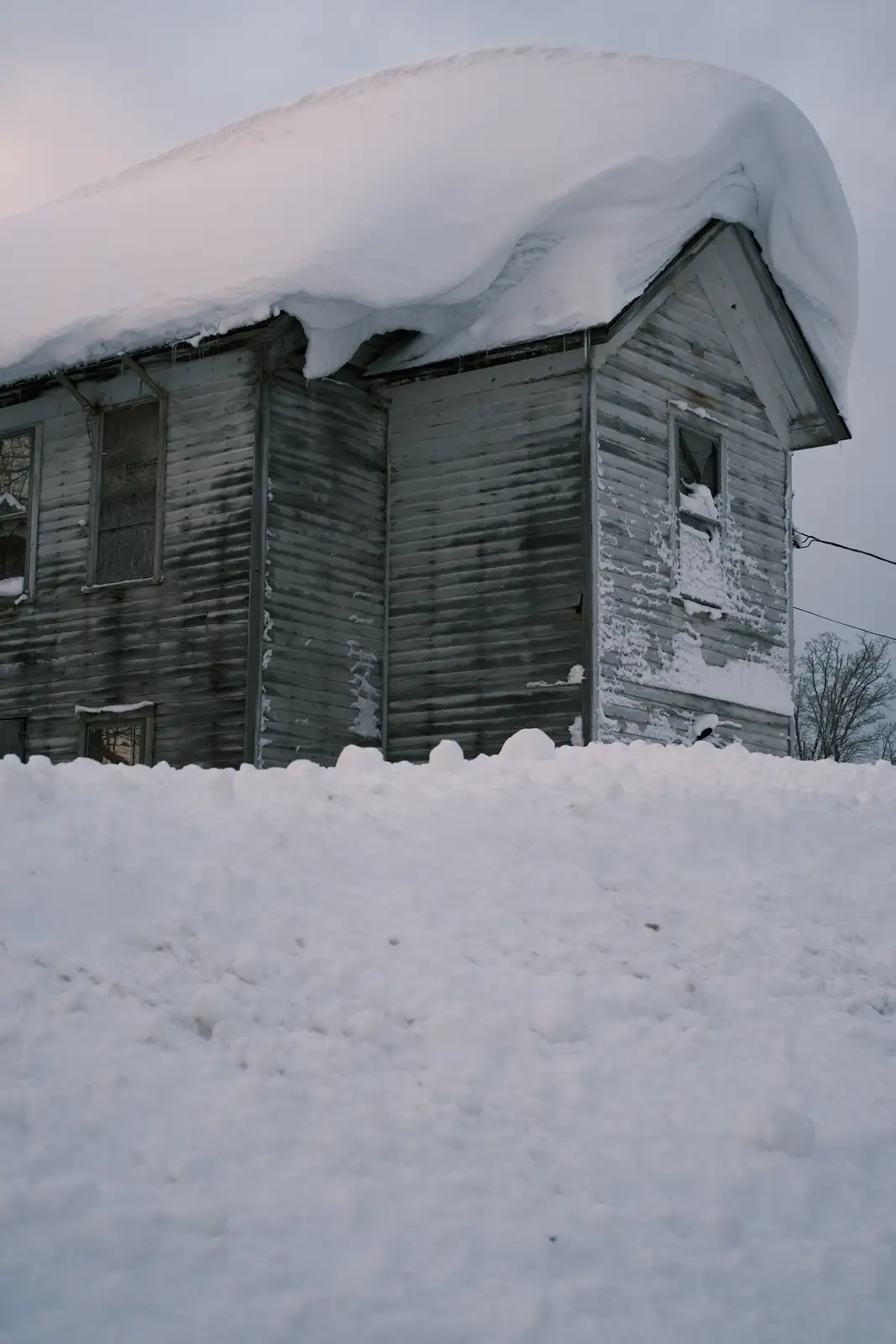 A weathered, gray wooden house is almost entirely buried under a thick blanket of snow, with snow piled high on its roof and…