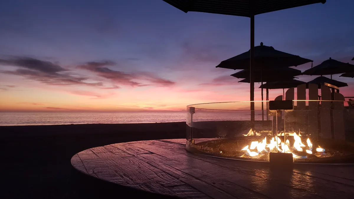 A serene twilight scene at a seaside resort features a glowing circular fire pit with flames dancing inside a glass…