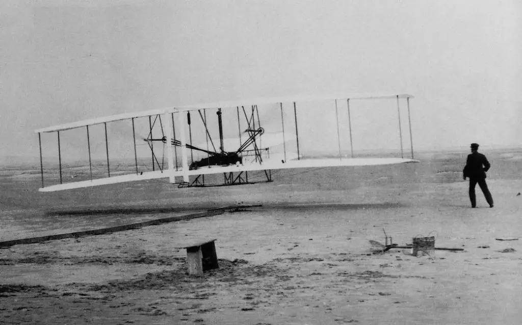 A black-and-white historical photograph shows the Wright brothers' 1903 Flyer biplane on a sandy beach, with a man in a dark…