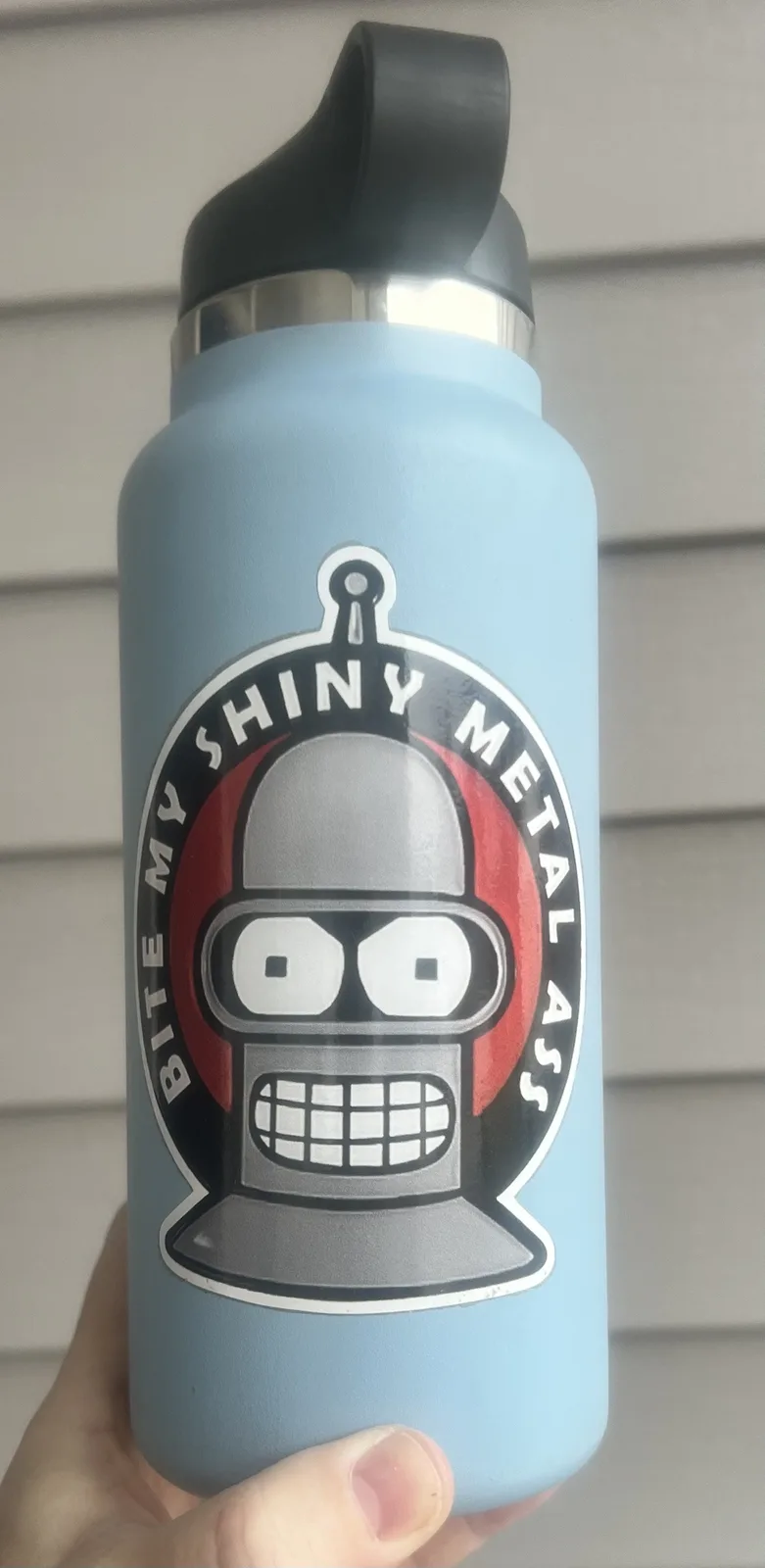 A light blue water bottle with a black lid is held in a hand, featuring a large sticker of the robot character Bender from…
