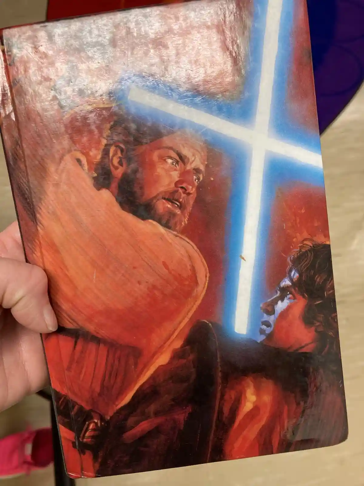 A hand holds a worn book with a painted cover depicting a bearded man in red robes looking upward toward a glowing blue…
