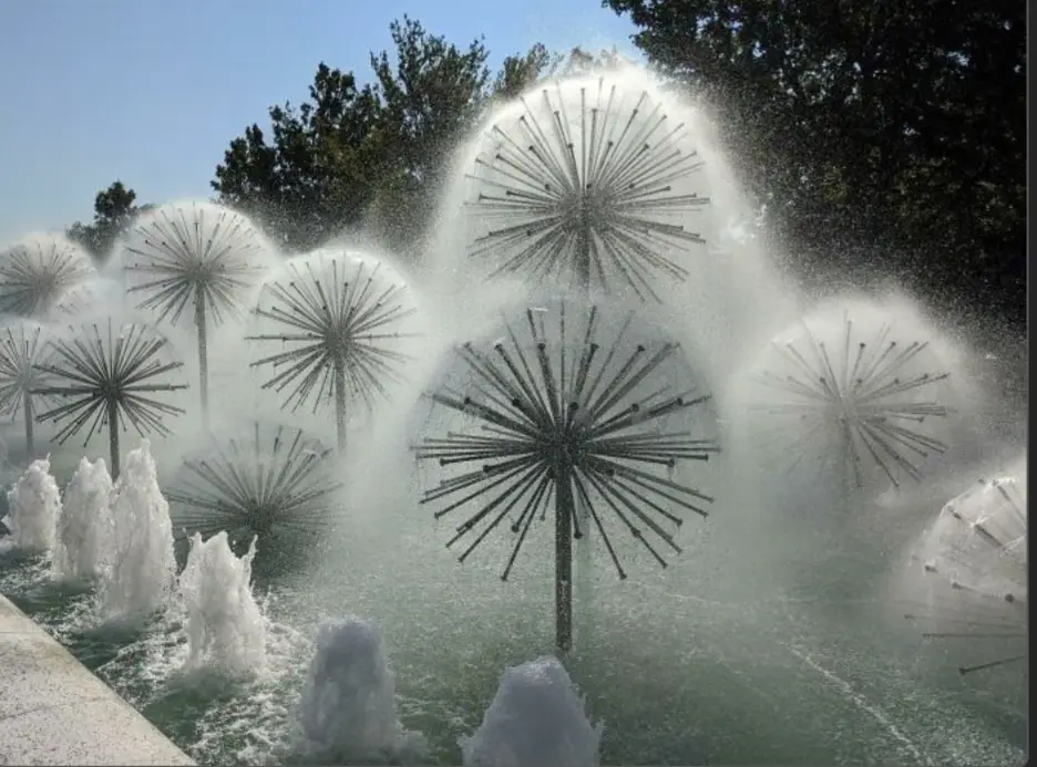 A photo of a fountain with multiple starburst-shaped water jets resembling dandelion clocks, set against a clear blue sky…