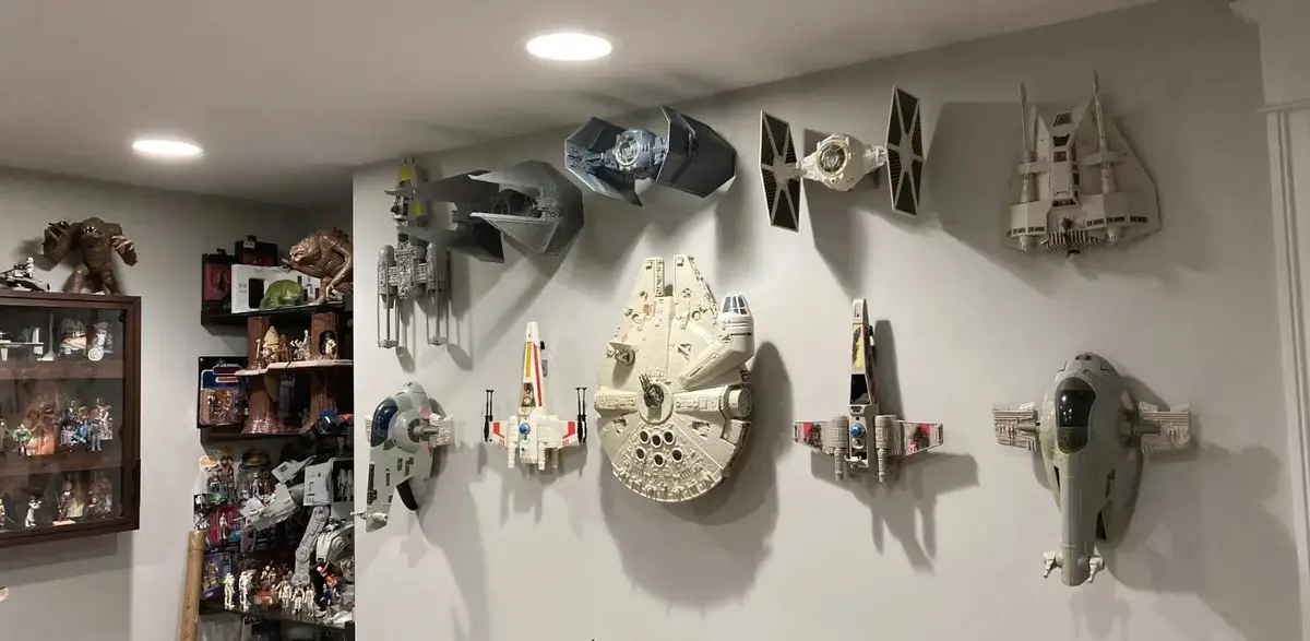 A room corner displays a large collection of Star Wars model ships mounted on a white wall, including the Millennium Falcon…
