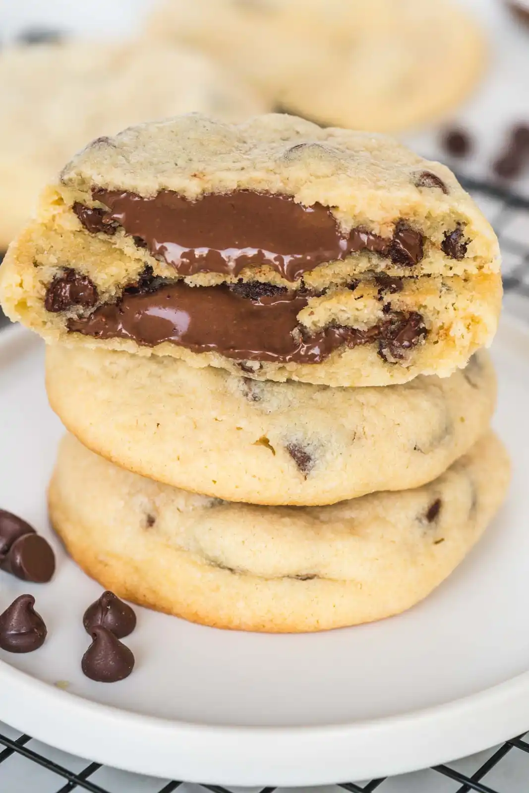 A stack of three golden-brown chocolate chip cookies sits on a white plate, with the top cookie broken open to reveal a…