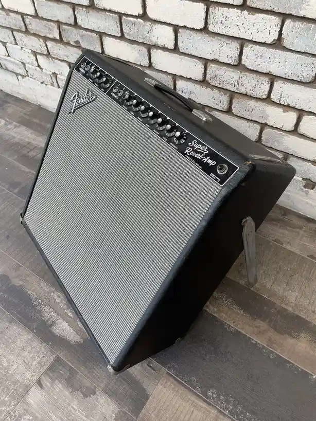 A vintage black Fender Super Reverb guitar amplifier sits at an angle on a gray wood floor against a white brick wall, its…