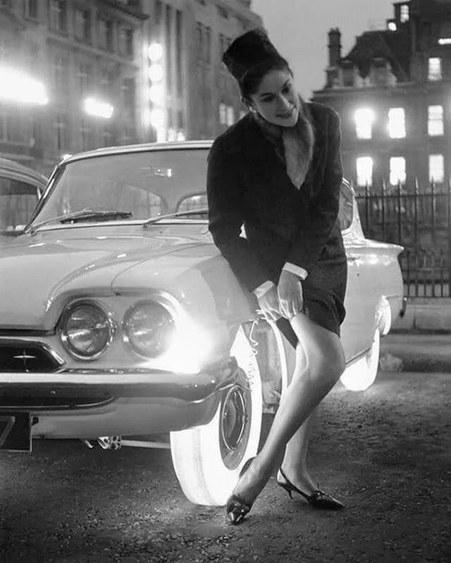 A black-and-white photo shows a woman in a 1950s-style outfit leaning against a vintage car at night, with the car's…