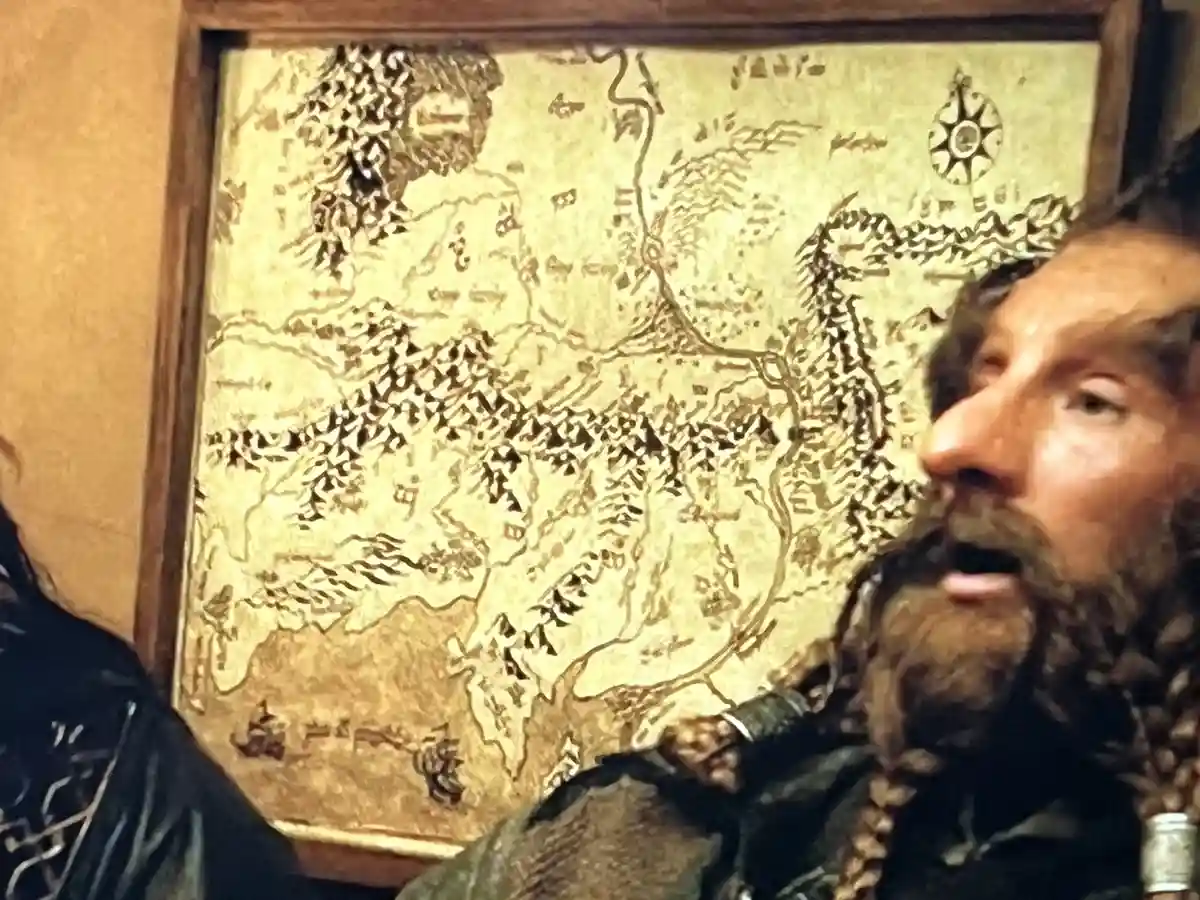 A man with a beard and braided hair looks at a framed, aged-looking fantasy map with a skeptical or unimpressed expression…