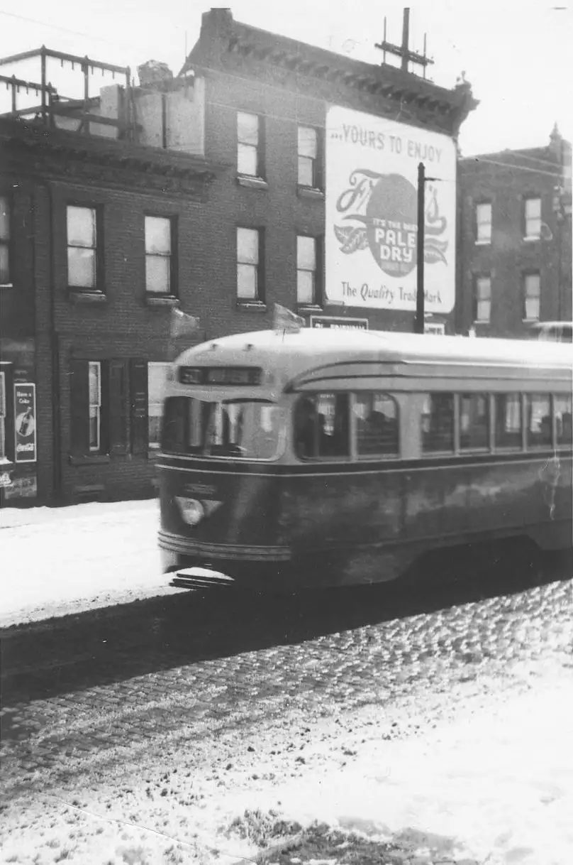 A black-and-white photo shows a vintage streetcar moving through a snowy urban street, passing brick buildings. A large…