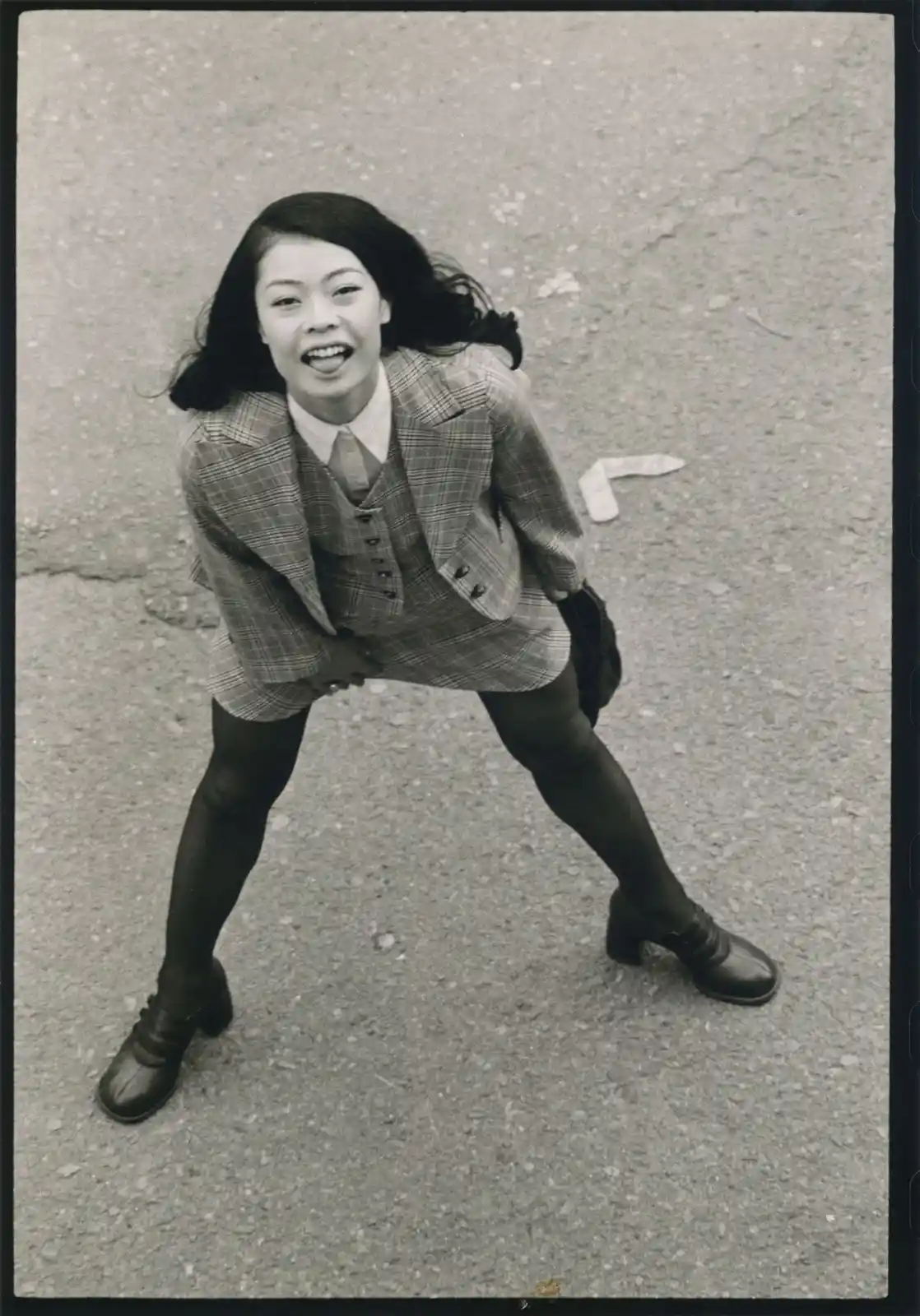 A black-and-white photo shows a young woman in a plaid school uniform, bending forward with her hands on her knees, looking…