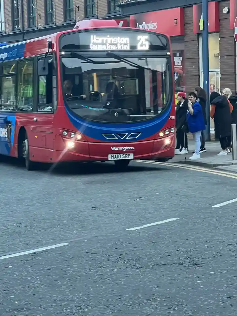 A red and blue bus with route 25 to Warrington is shown driving down a street, with several people waiting at a bus stop…