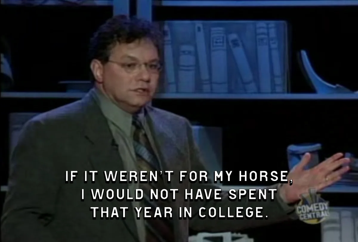 A man in a suit and glasses is speaking on a talk show set with bookshelves in the background. White text overlaid on the…
