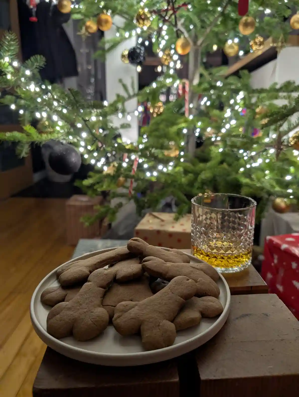 A plate of gingerbread cookies shaped like bones sits next to a glass of whiskey on a wooden table, with a decorated…