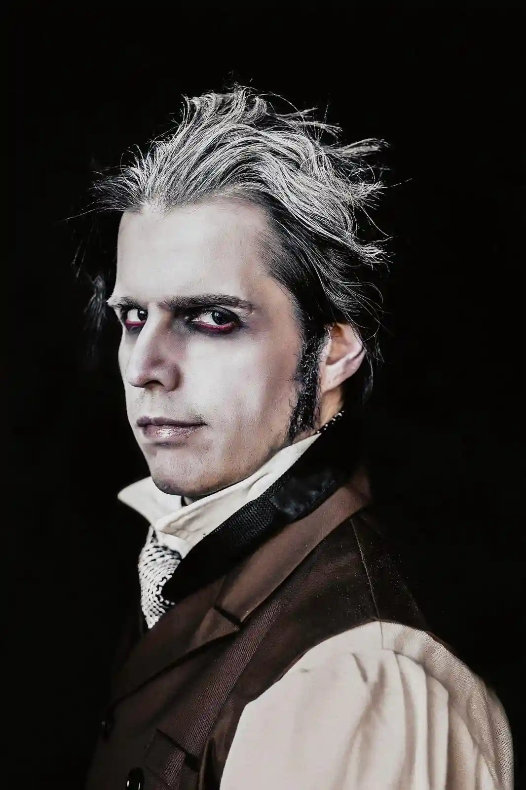 A man with pale skin, dark eye makeup, and red-tinged eyes stares intensely at the camera against a black background. He…
