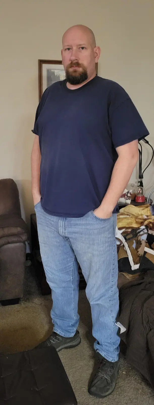 A bald man with a full beard stands indoors wearing a blue t-shirt and jeans, hands in his pockets, looking directly at the…