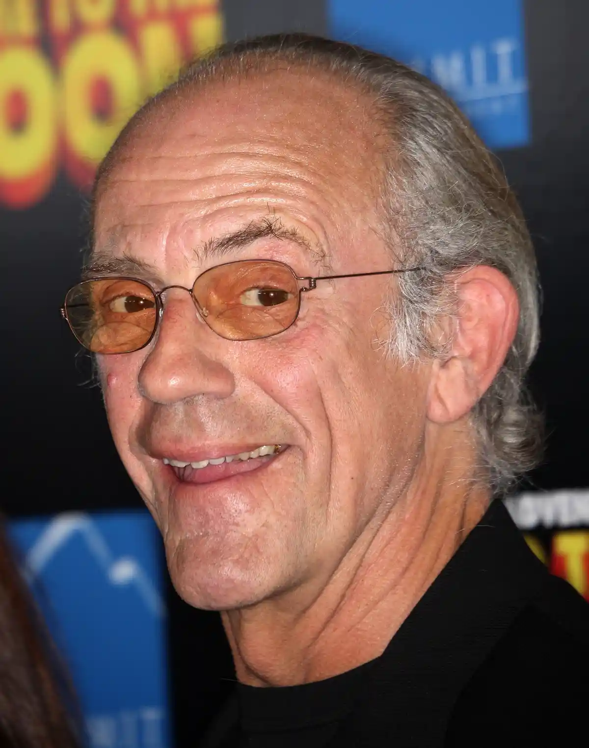 A close-up photo of an older man with glasses and gray hair smiling slightly while wearing a black shirt. Behind him…