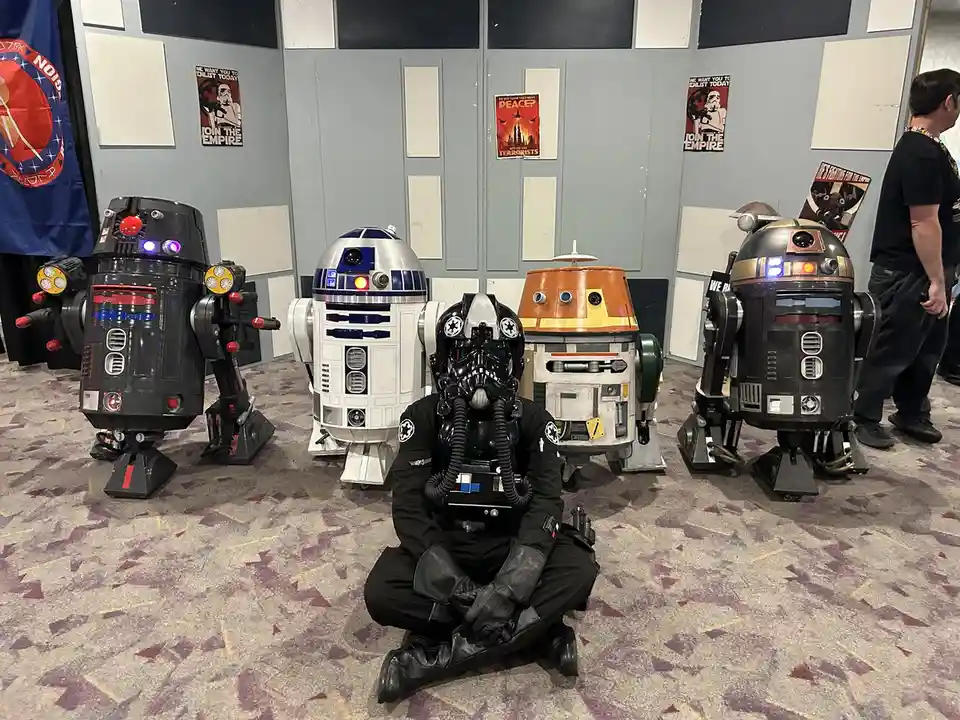 A person in a black stormtrooper costume sits cross-legged on a patterned carpet, surrounded by four Star Wars droids…