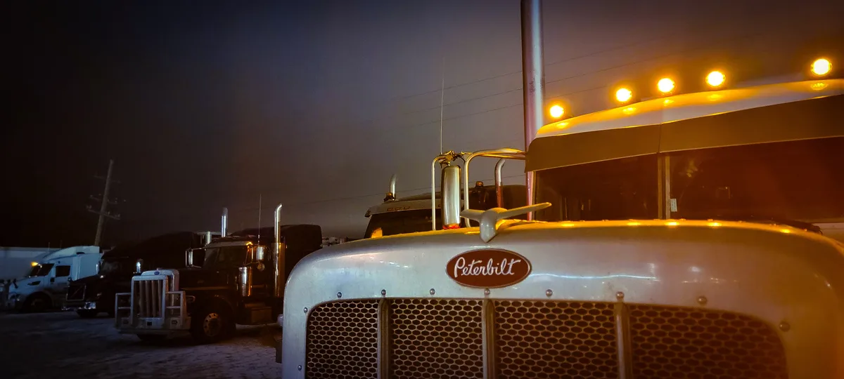 A close-up of a white Peterbilt semi-truck's front grille and hood, illuminated by its own amber roof lights at night, with…