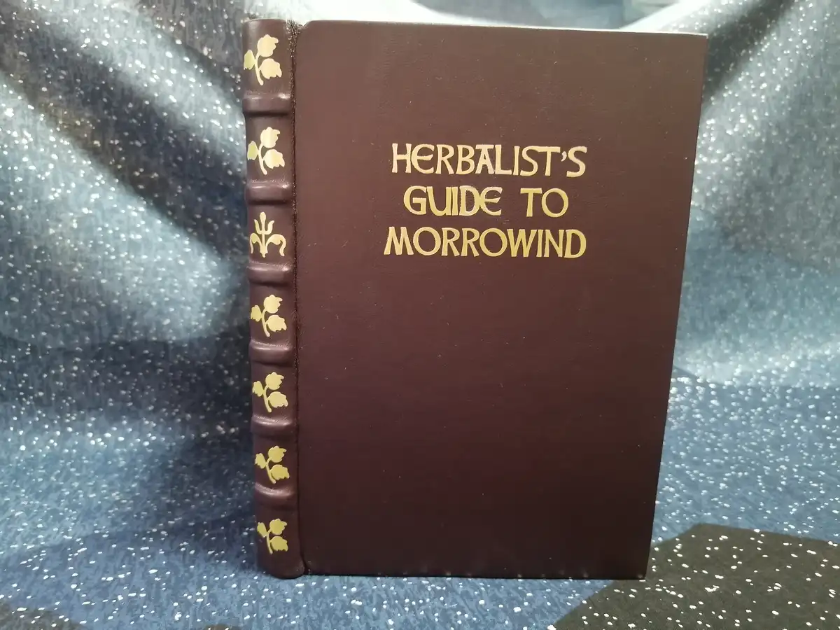 A brown leather-bound book with gold embossed lettering reading 'HERBALIST'S GUIDE TO MORROWIND' and gold leaf motifs on its…