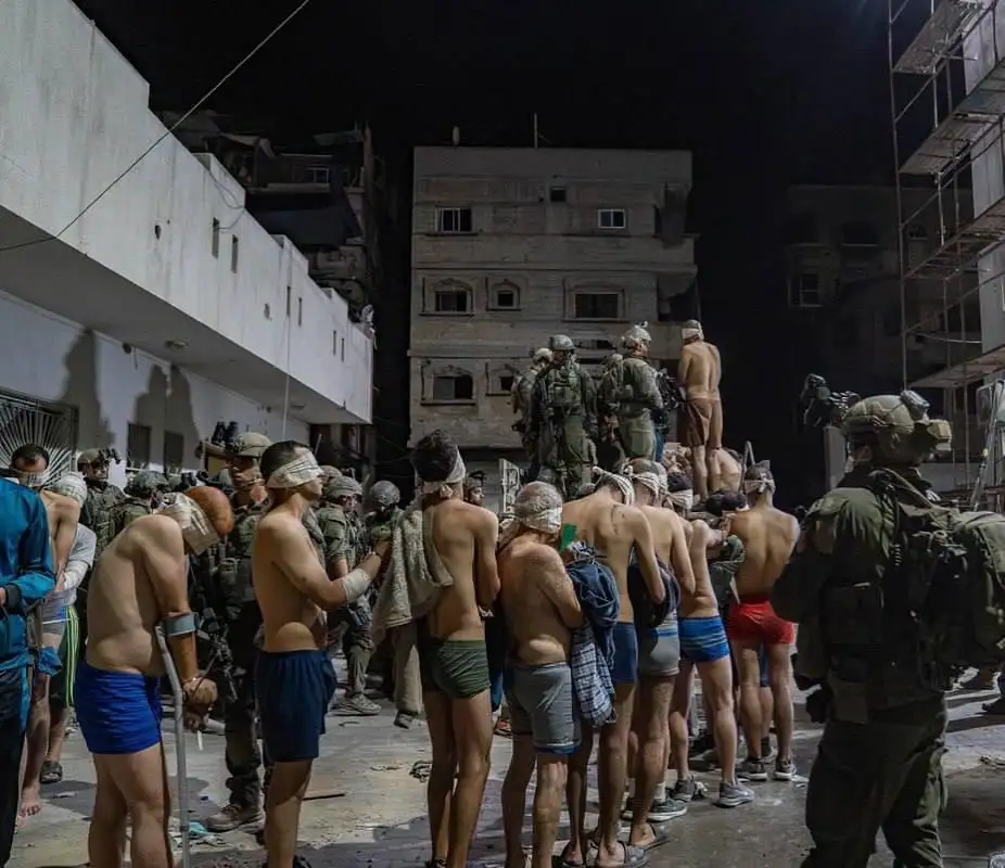 A nighttime photo shows a group of shirtless men in underwear and some with bandages on their heads, standing in a line…