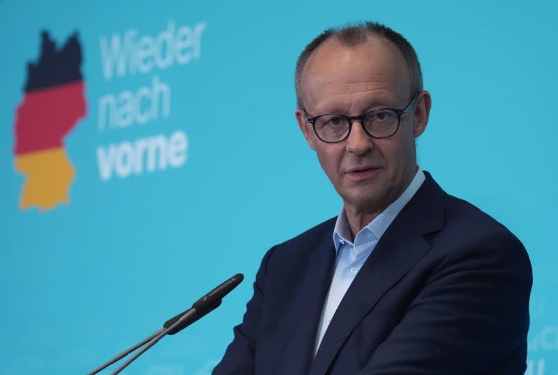 A man in glasses and a suit stands at a podium in front of a blue backdrop featuring a German map outline colored with the…