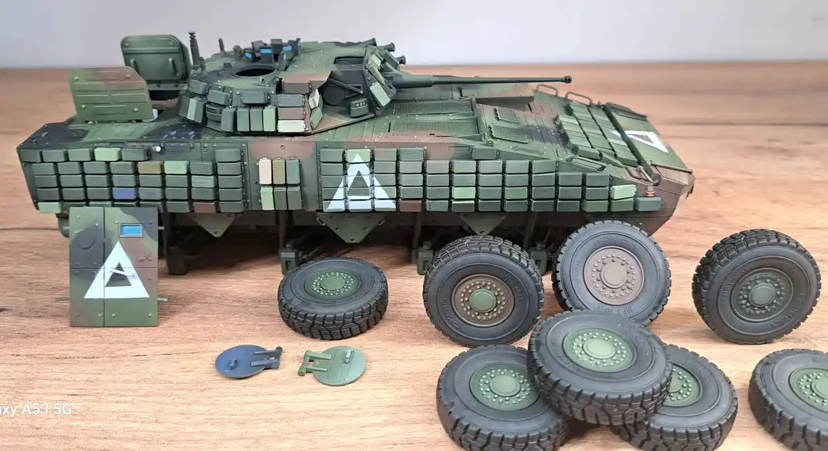 A detailed green military tank model is displayed on a wooden surface, surrounded by its unassembled parts including…