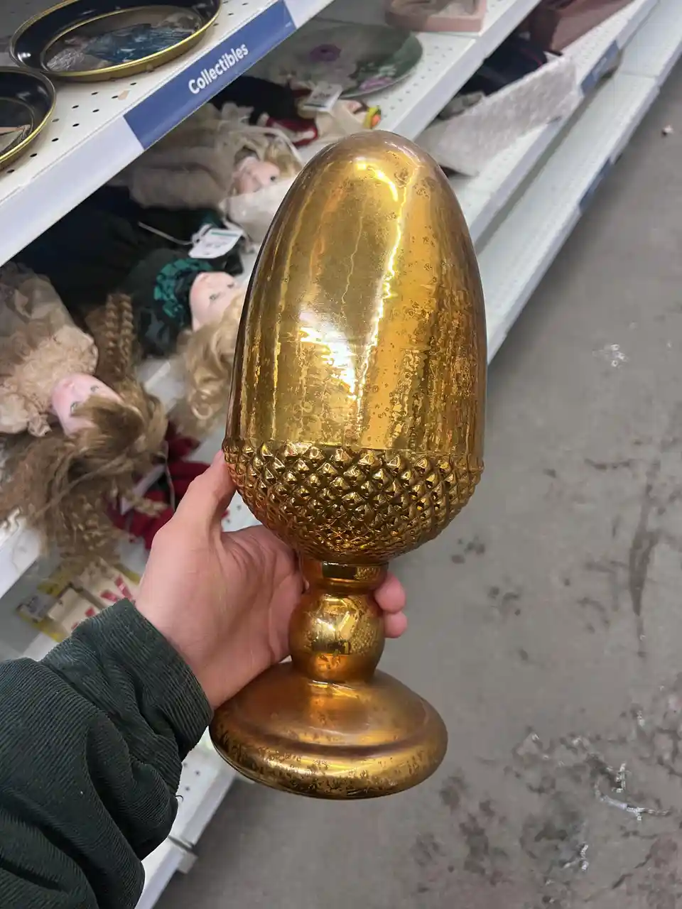 A hand holds a large, ornate golden object shaped like a pinecone with a textured base and stem, in front of a store shelf…