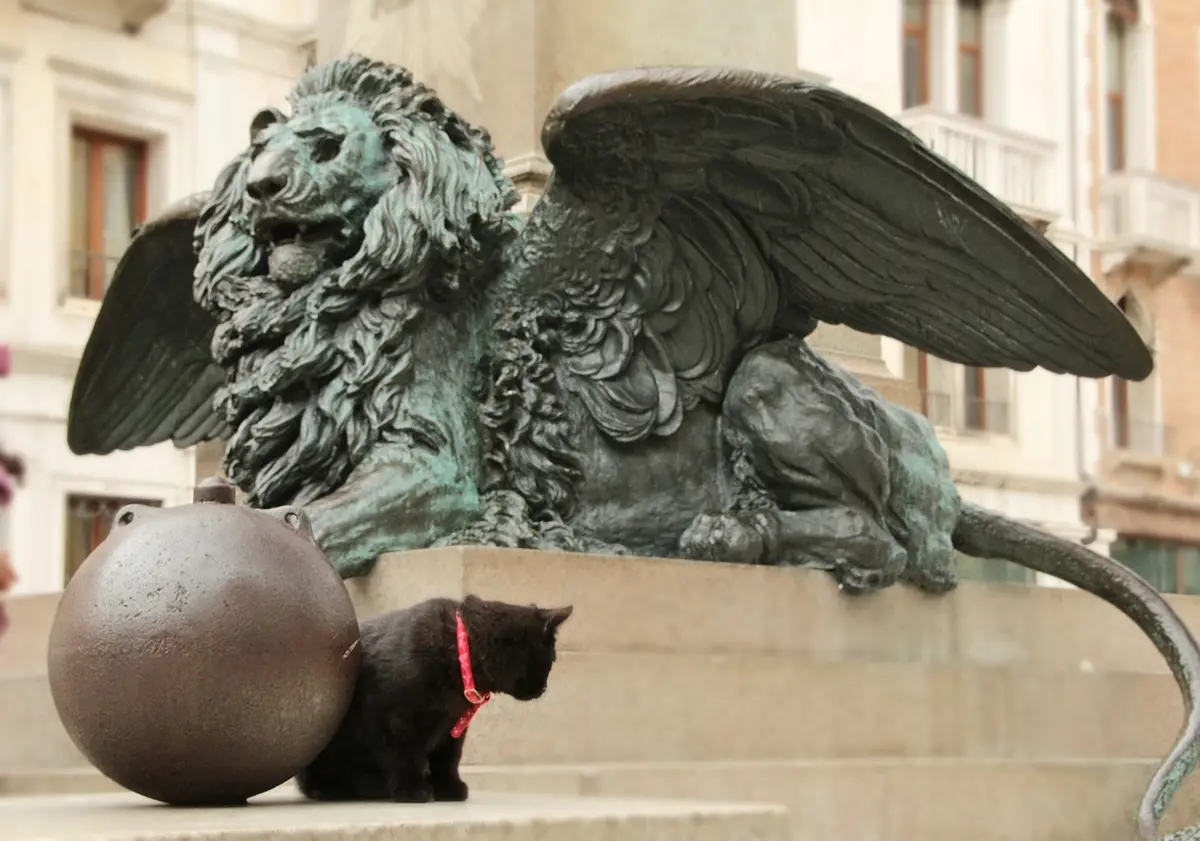A black cat with a red collar sits beside a large cannonball at the base of a bronze winged lion statue. The statue, showing…