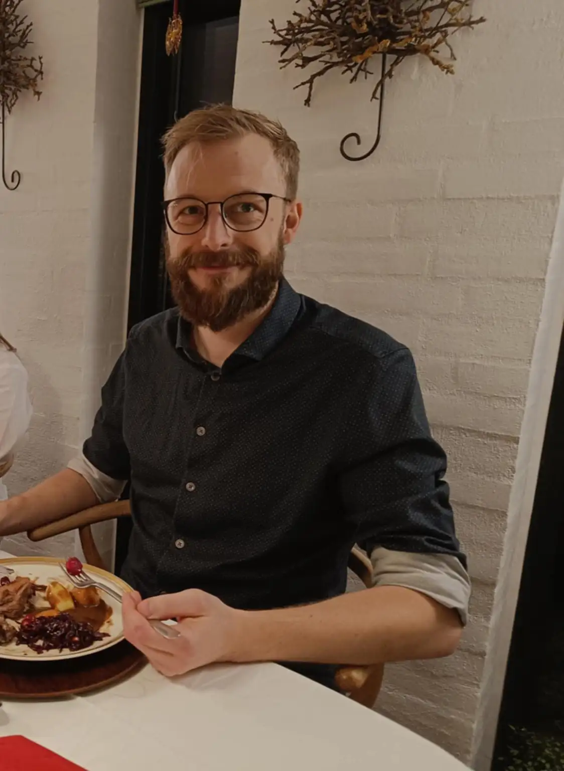 A man with glasses and a beard sits at a restaurant table with a partially eaten plate of food, smiling at the camera. The…