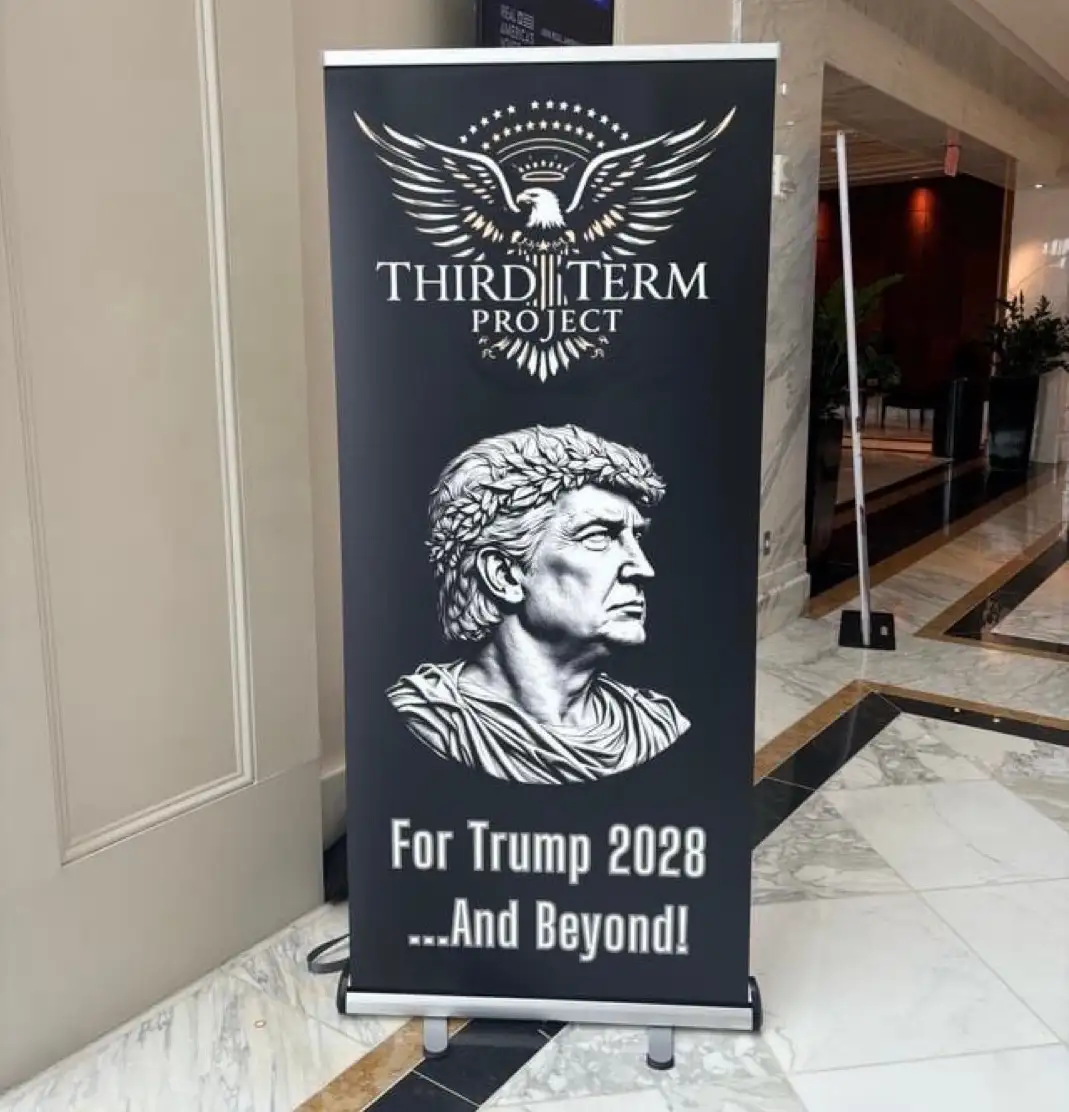 A black banner stand in a marble-floored lobby displays a stylized Roman bust of Donald Trump wearing a laurel wreath, under…